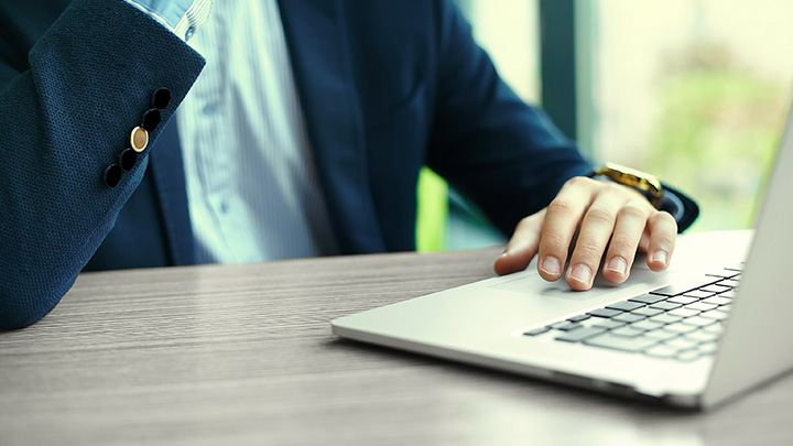 Close up of a man's hand on a laptop. Man is wearing a dark blue suit jacket and light blue shirt