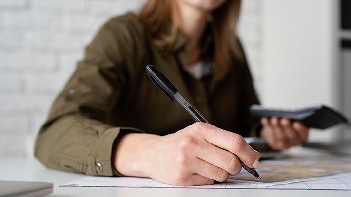 A close up of a woman's hand holding a pen, writing on documents on a desk, She is holding a calculator in her other hand